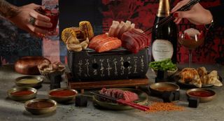 Two hands reaching from either side of the image above a spread of meat, fish, vegetables, bowls of dipping sauce and a Hibachi grill