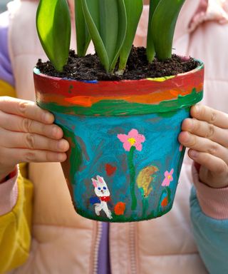 Two small hands holiding a painted flowerpot with a bunny and flower painted onto it, and green shoots in the brown soil, with a pink pastel puffer jacket in the background
