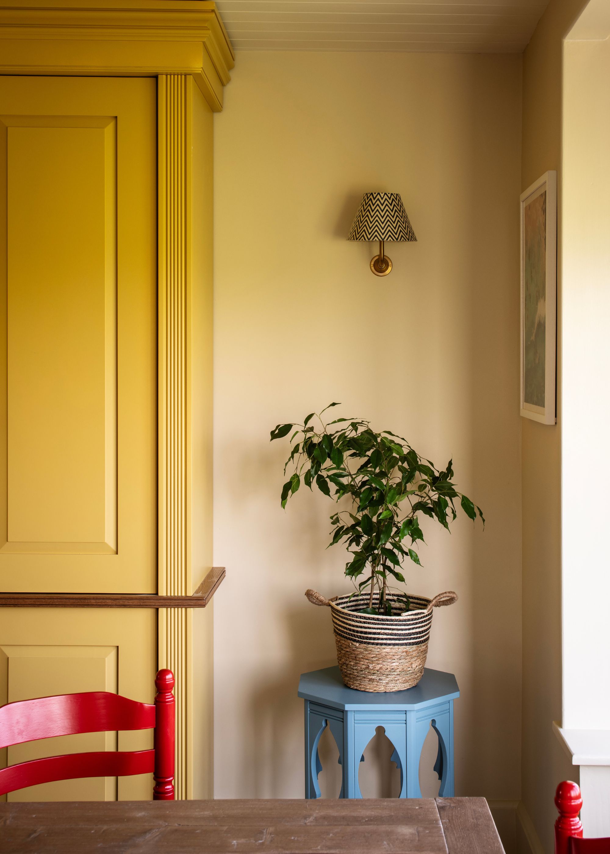 A dining room with yellow walls and a yellow cabinet, a bright red chair, and a blue side table with a plant.