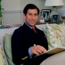 Prince Charles wearing khakis and a black blazer sitting on a sofa with green floral pillows in his living room with a table and lamp behind him