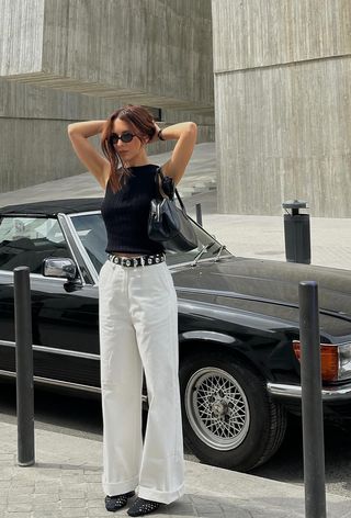 The woven shoe trend is shown in a street-style photo of a woman standing outside by a black car wearing black sunglasses, a black top, a black studded belt, white pants, a black shoulder bag, and black woven leather flats