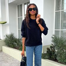 Influencer @lornasymphony stands outside wears a navy jumper with mid-wash blue jeans and white open-toe heels.
