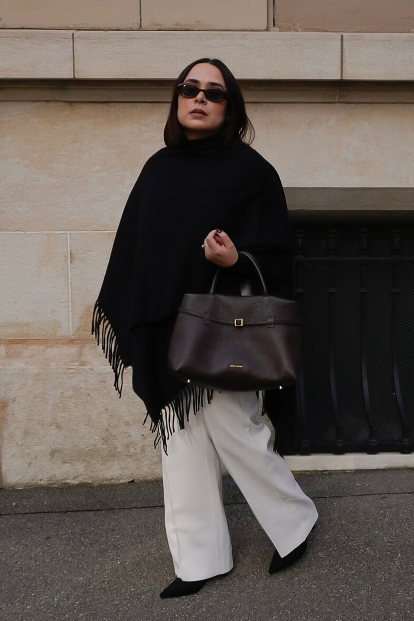A woman standing on the sidewalk wearing brown sunglasses with a black cape, white wide-leg trousers, black pointed heels, and a brown handbag.