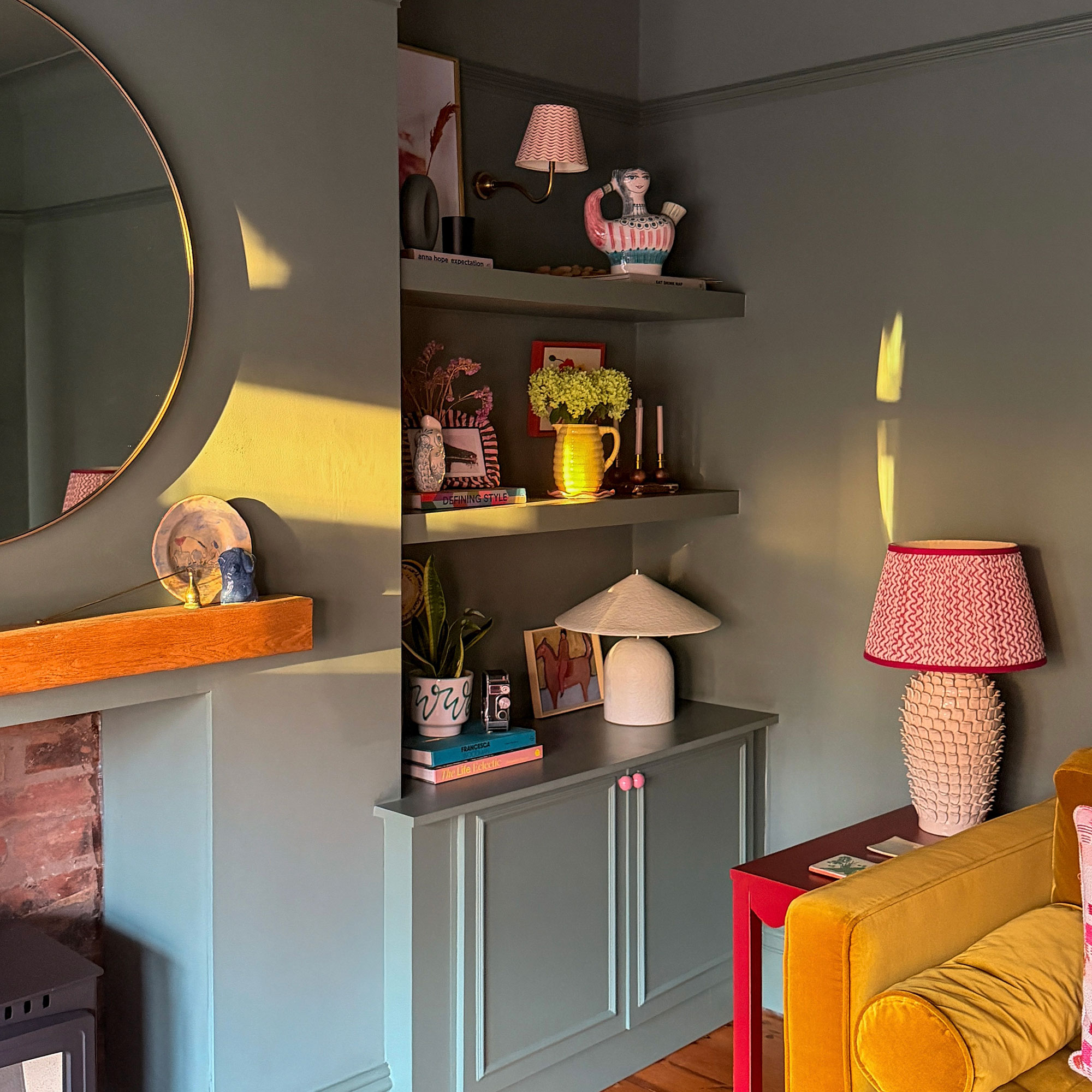 Green living room with built in cupboards and shelving filled with vases, candles, books and art beside mustard sofa, red side table and white ceramic table lamp with red and white shade