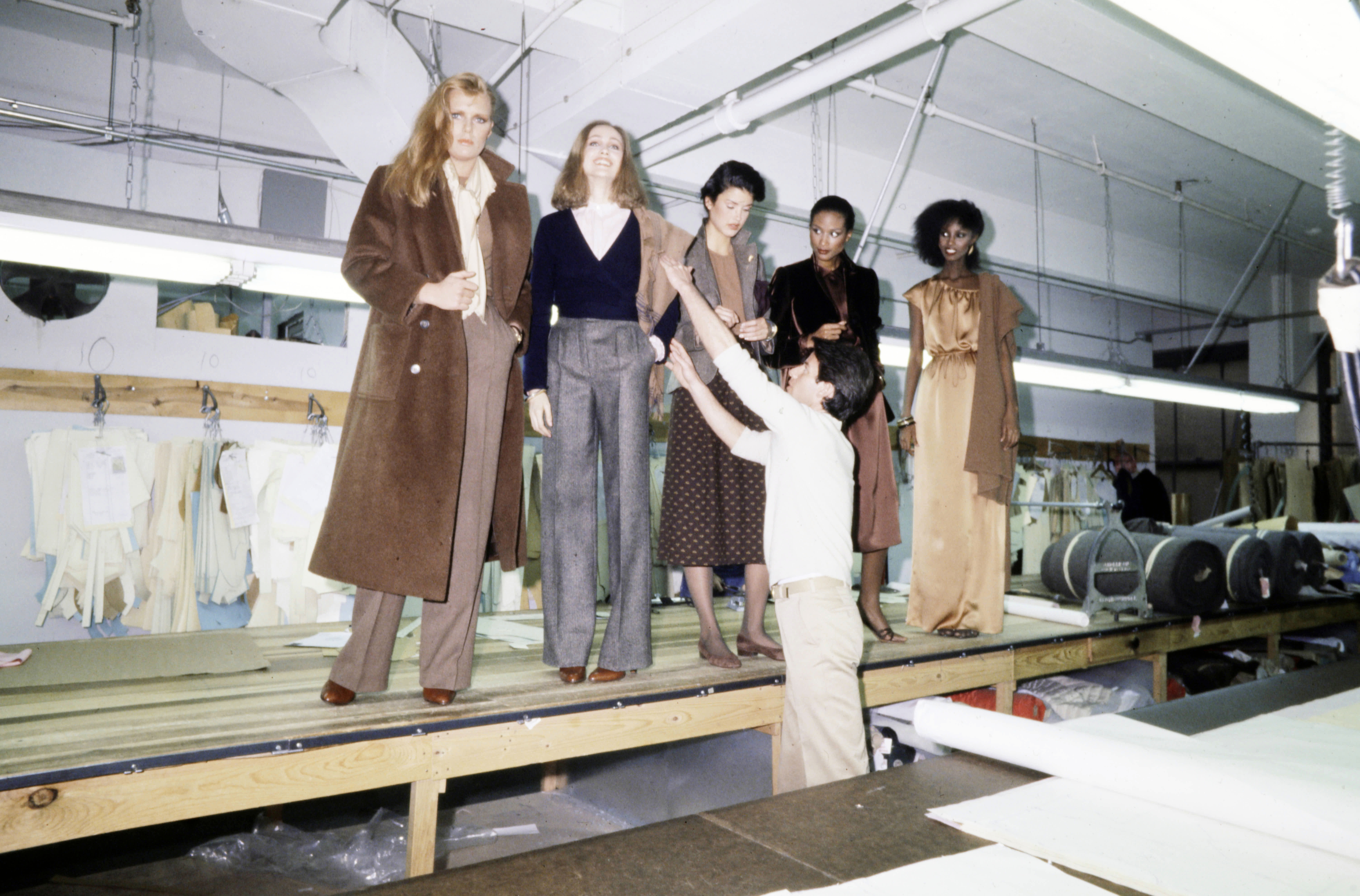 A young male fashion designer dressed in beige clothing perfects the winter look of a series of models standing in his fashion workshop.