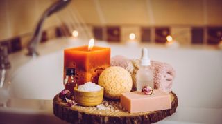 Home spa products on wooden tray: bar of soap, bath bomb, aroma bath salt, essential and massage oils, candle burning, rolled towel inside bathroom by tub running water