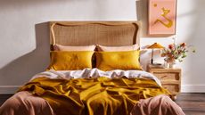 Yellow, orange, and white bed sheets on a wooden bed frame beside a wood nightstand and orange wall art against a white wall. 