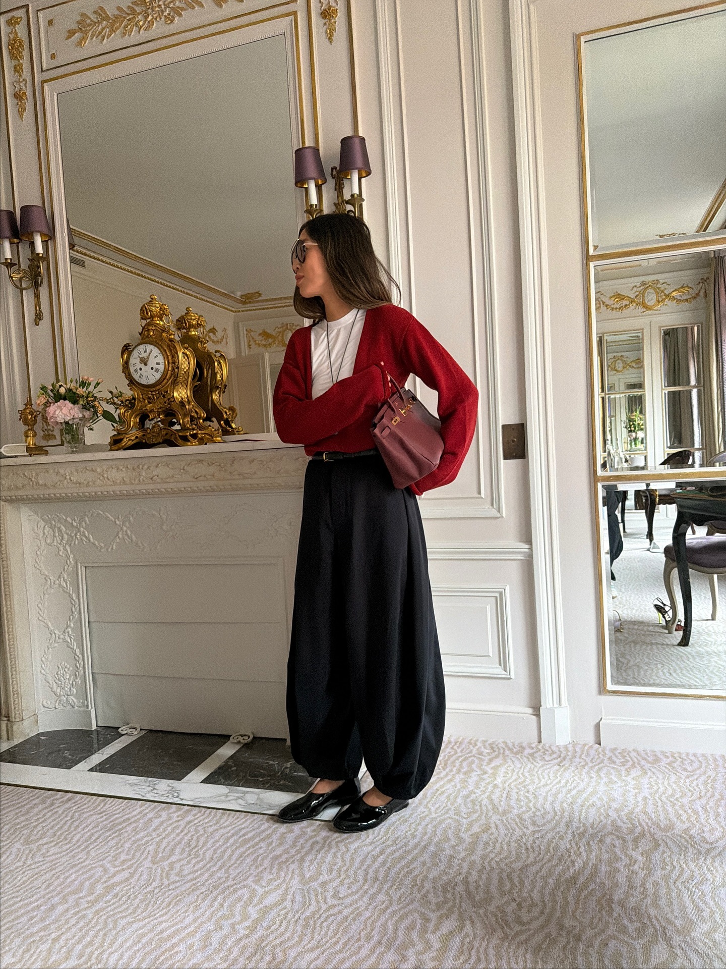 Aimee wears a red sweater, white tee, black balloon pant, patent leather flats, and a leather burgundy bag.