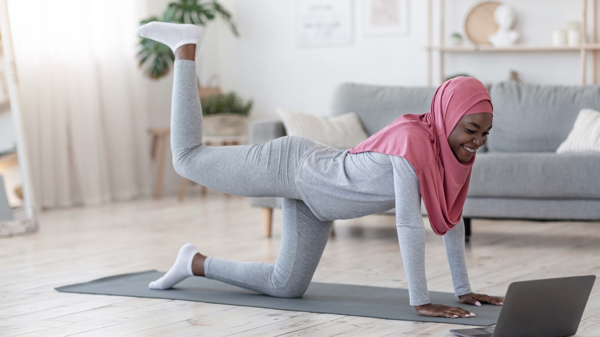 woman dressed in light grey with a pink hijab on all fours with one leg kicking up behind her. she's on a grey exercise mat on wooden floor with a laptop on the floor in front of her. there's a grey sofa behind her.