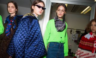 Model wears narrow sunglasses with a navy quilted jacket. Another wears a neon green jumper