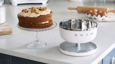 Smeg digital scales in white on a marble countertop with a carrot cake on a stand beside it