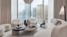 beige living room with curved white sofe and beige chubby armchairs and a curved ottoman, with marble coffee table styled with vases and beige curtains around the room and floor to ceiling windows with city outlook