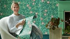 Image of Ruther Mottershead from Little Greene sat on a chair holding green patterned wallpaper. 
