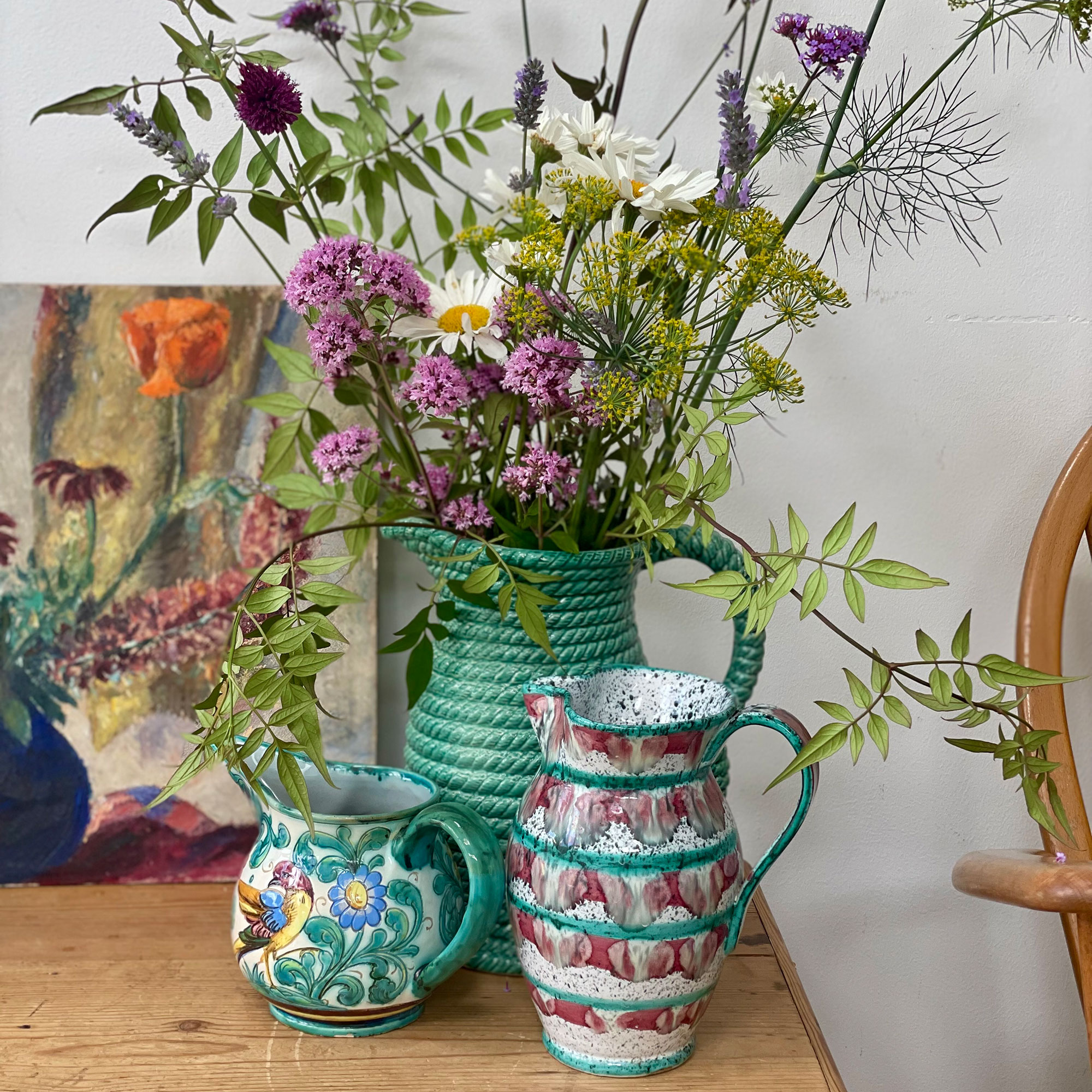 Three ceramic jugs, one with bunch of flowers, in front of piece of art