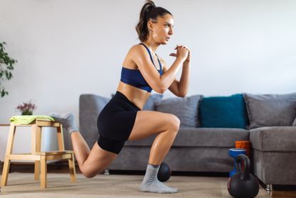 Woman exercising at home next to a couch. She is midway through performing a Bulgarian split squat, with her rear foot elevated on a stool and both knees bent to 90°