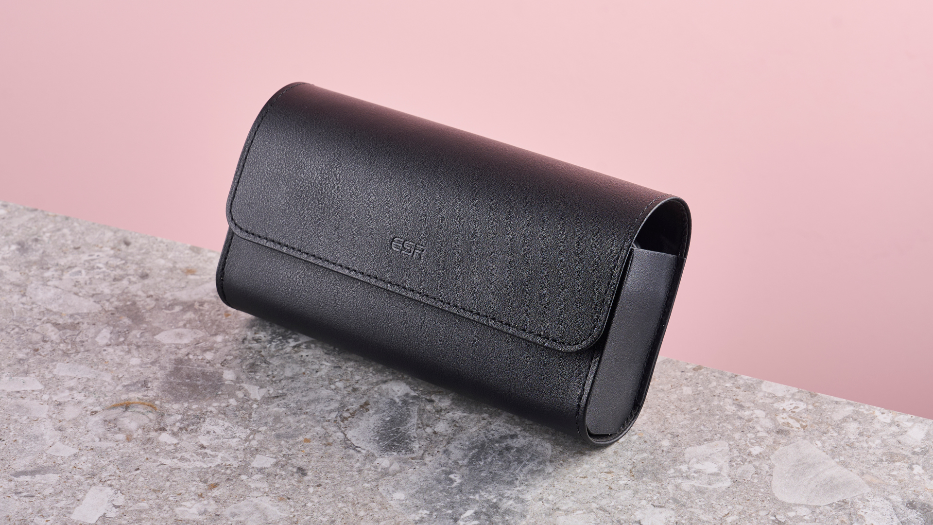 The black leather-effect travel case sits on a stone-effect surface in front of a pink background.