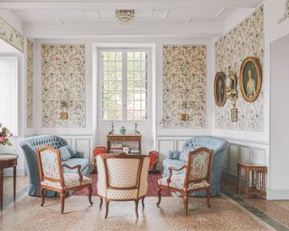 French house living room with floral wallpaper, gold framed portraits, matching pale blue couches, antique armchairs, tiled floor, rug, wall lights, side tables