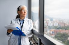 A female doctor holding a clipboard at the hospital and smiling while looking through the window.
