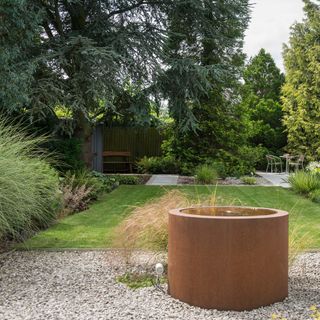 a corten steel round water feature beside a large lawn area and bistro seating on the other side of the garden