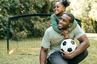 A black father and daughter are in a garden with a goal post behind them, they are playing football.