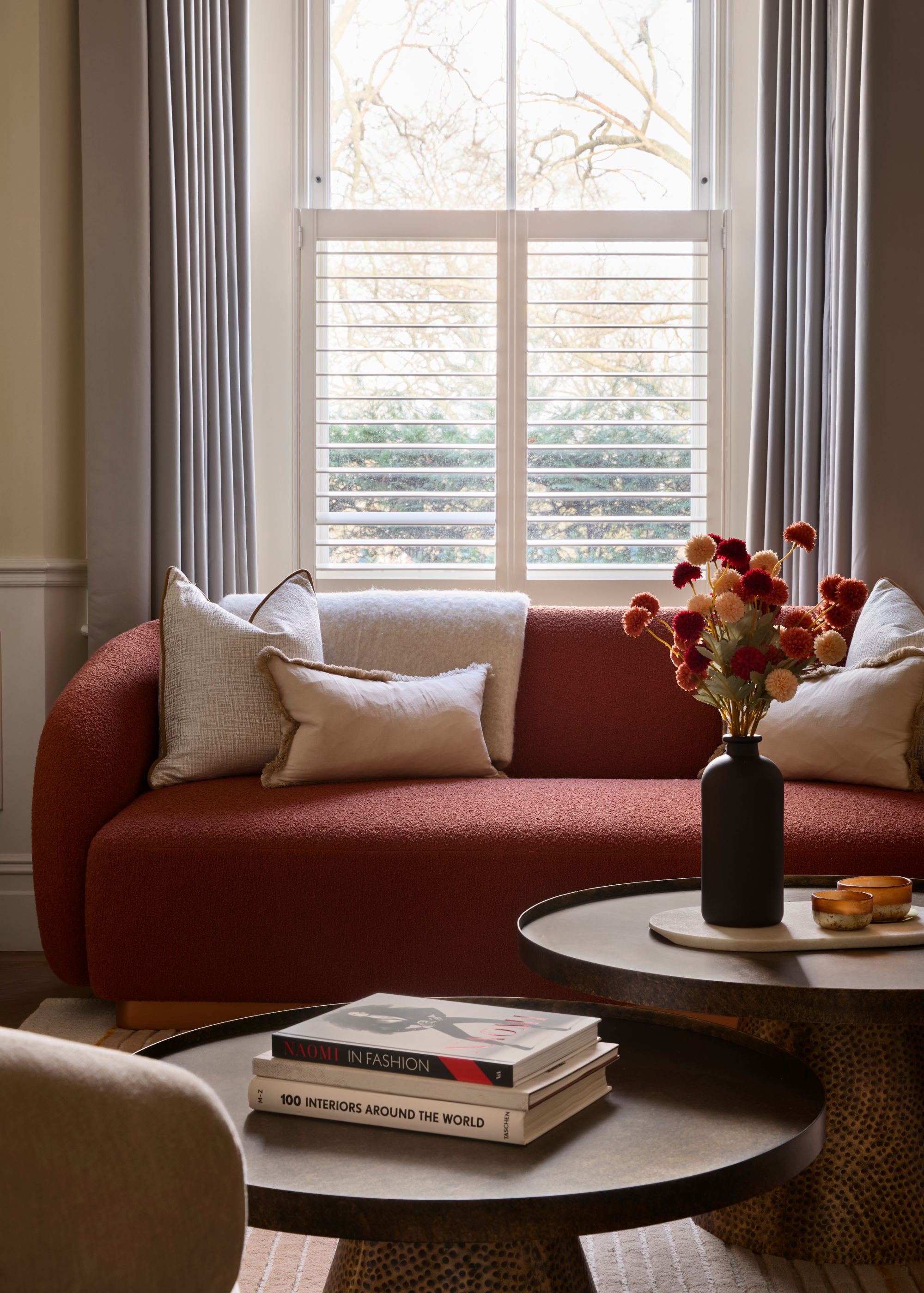 Two round nesting coffee tables are styled with books and decorative objects as well as a dark vase with flowers next to a red sofa in front of a window