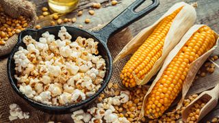 Popcorn ligger i en liten gjutjärnspanna bredvid ett gäng kärnor och majskolvar på ett bord.