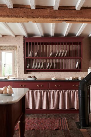 dark red kitchen with large plate rack on wall, fabric skirt, brass handles, black countertop, wooden floorboards red vintage rug, pale pink walls, beams
