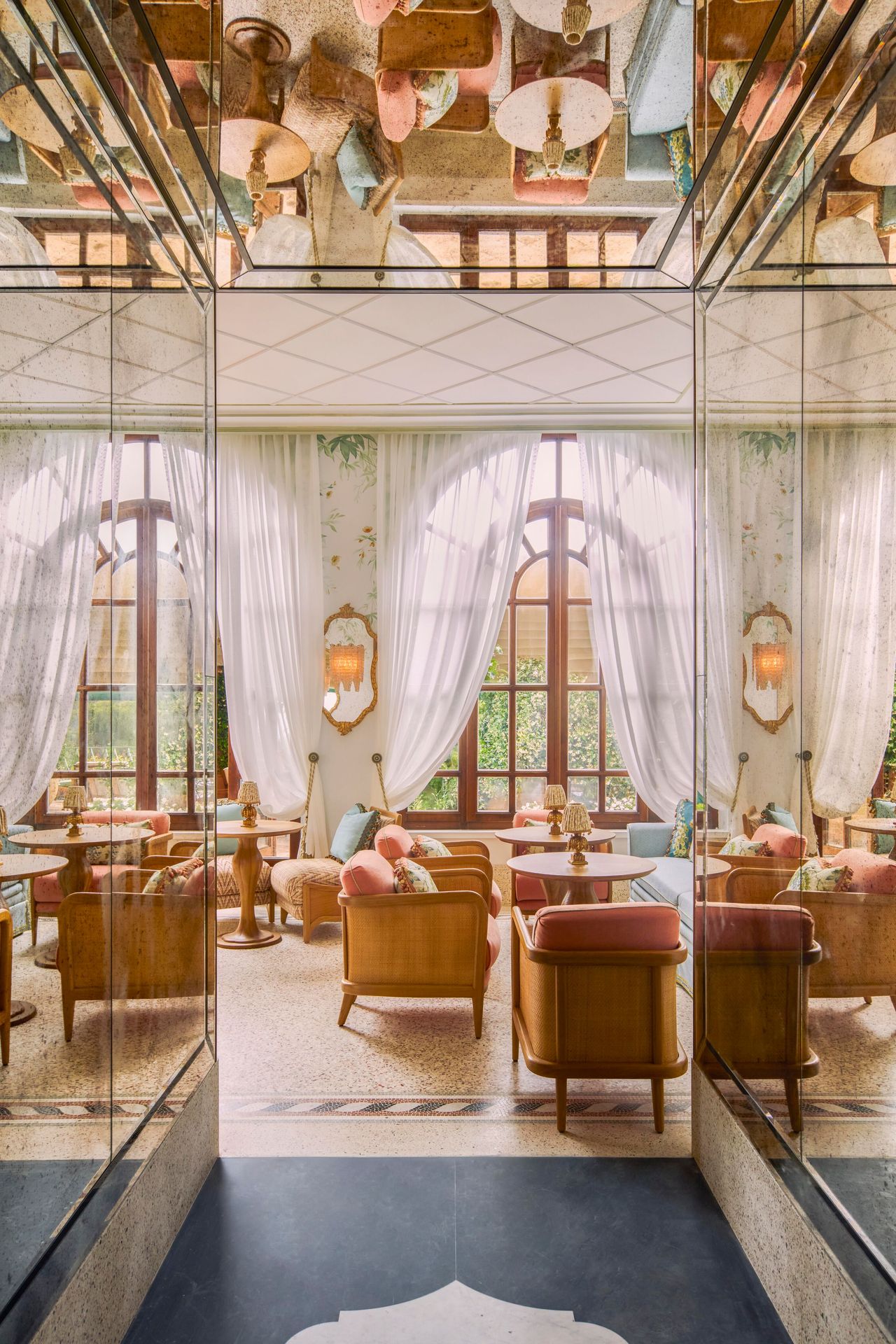 A mirrored corridor opens up into the seating area of a bar with large windows and pale wooden chairs and tables