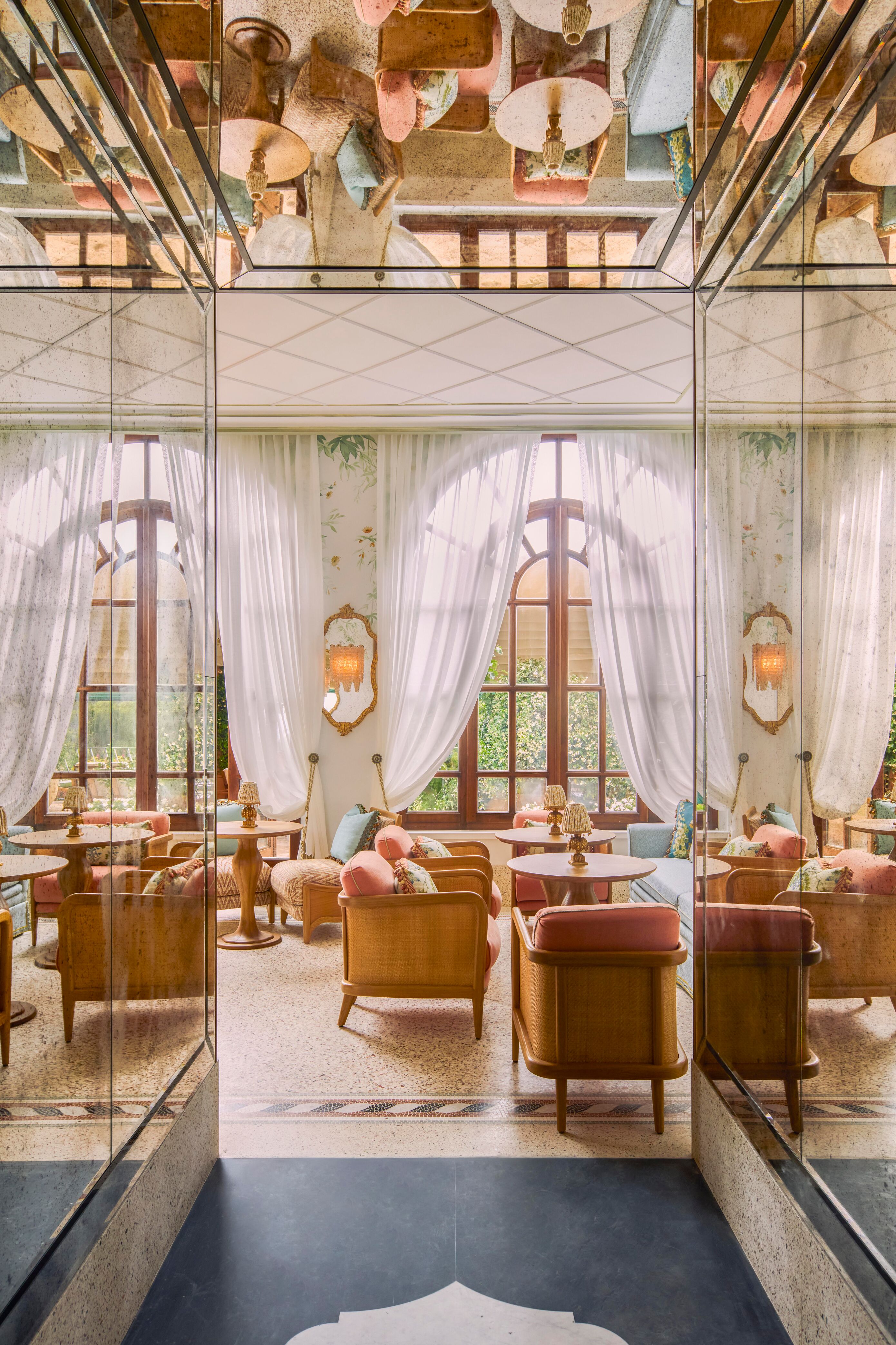 A mirrored corridor opens up into the seating area of a bar with large windows and pale wooden chairs and tables