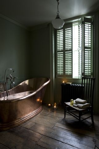 An example of green bathroom ideas showing a green bathroom with green walls and green painted shutters and a copper bath with wooden original floorboards