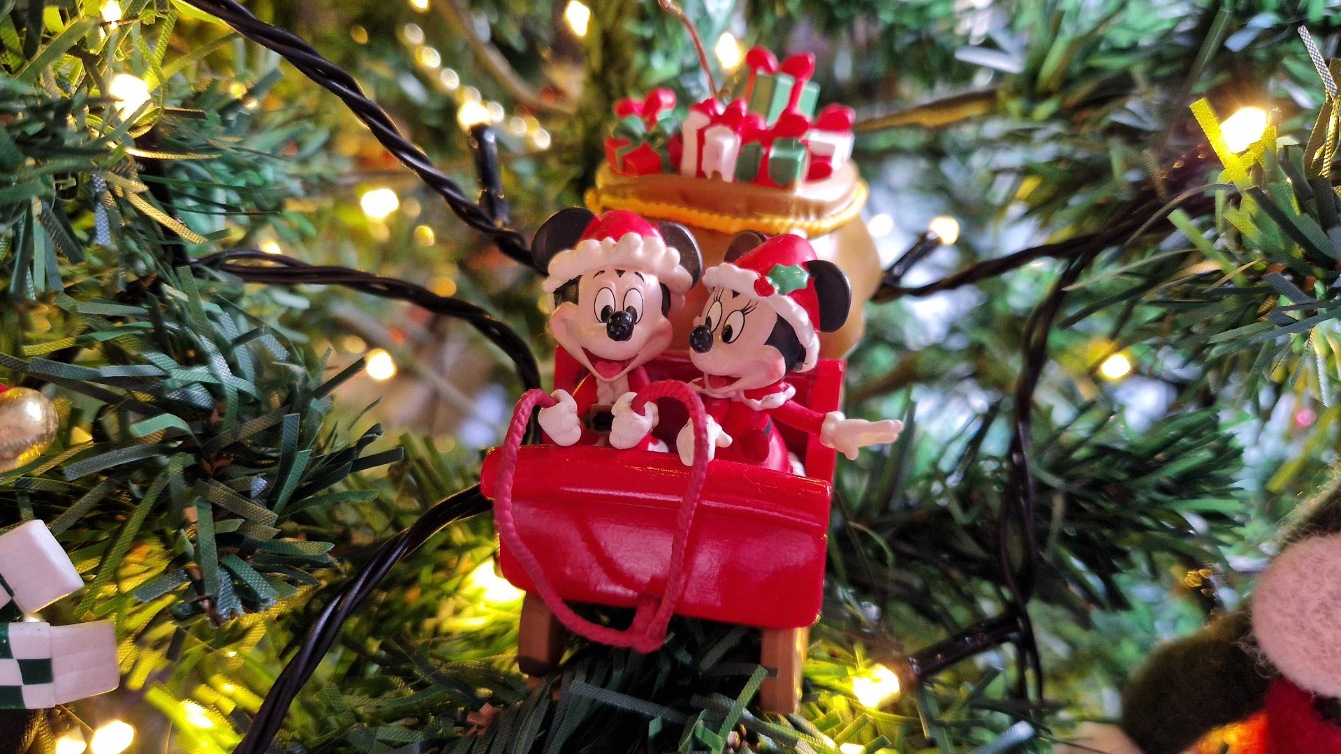 Mickey and Minnie Mouse wearing Santa outfits in a red sleigh packed with presents, sat amongst Christmas tree foliage with twinkling lights around them