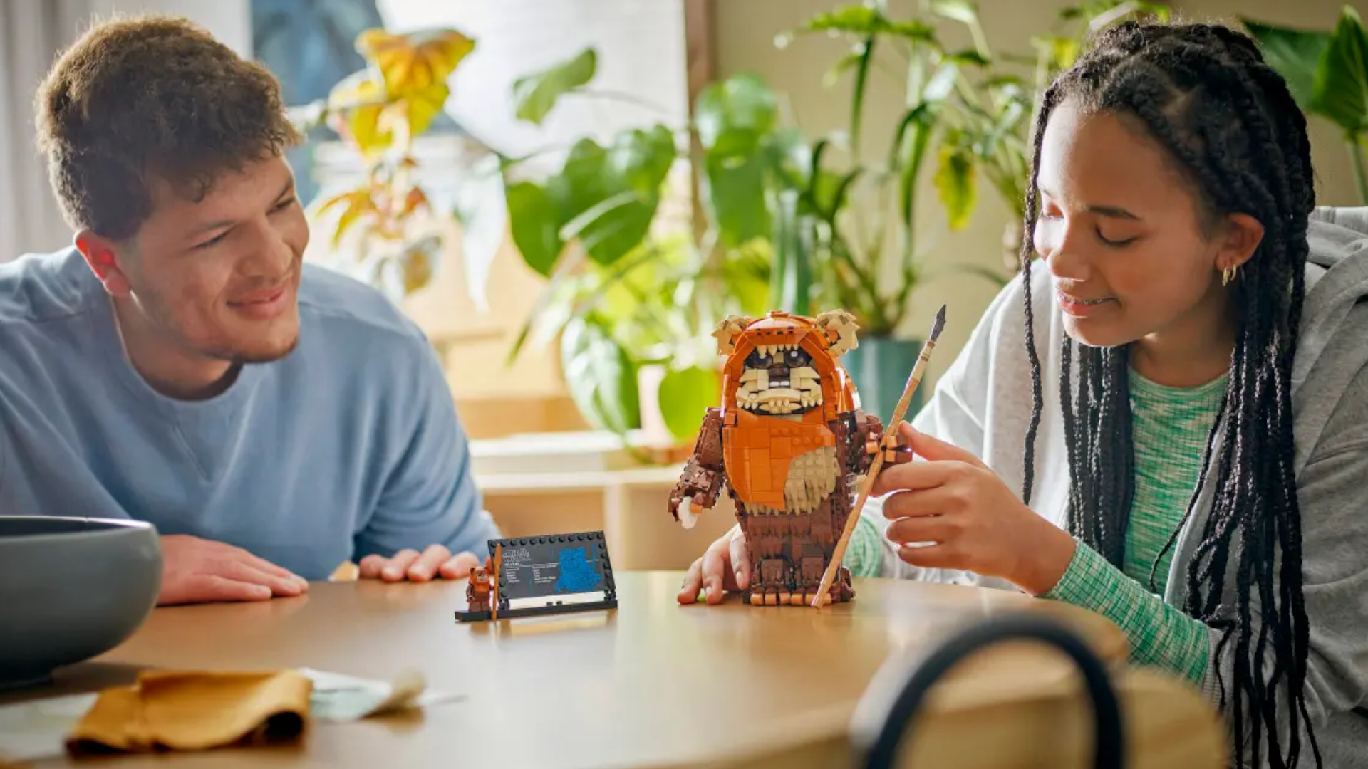 A young woman poses the Lego Wicket the Ewok model as it stands on a plain table, while a man looks on with a smile