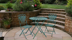 Teal wrought iron outdoor bistro table and chairs sitting on a stone patio surrounded by brick planters filled with greenery
