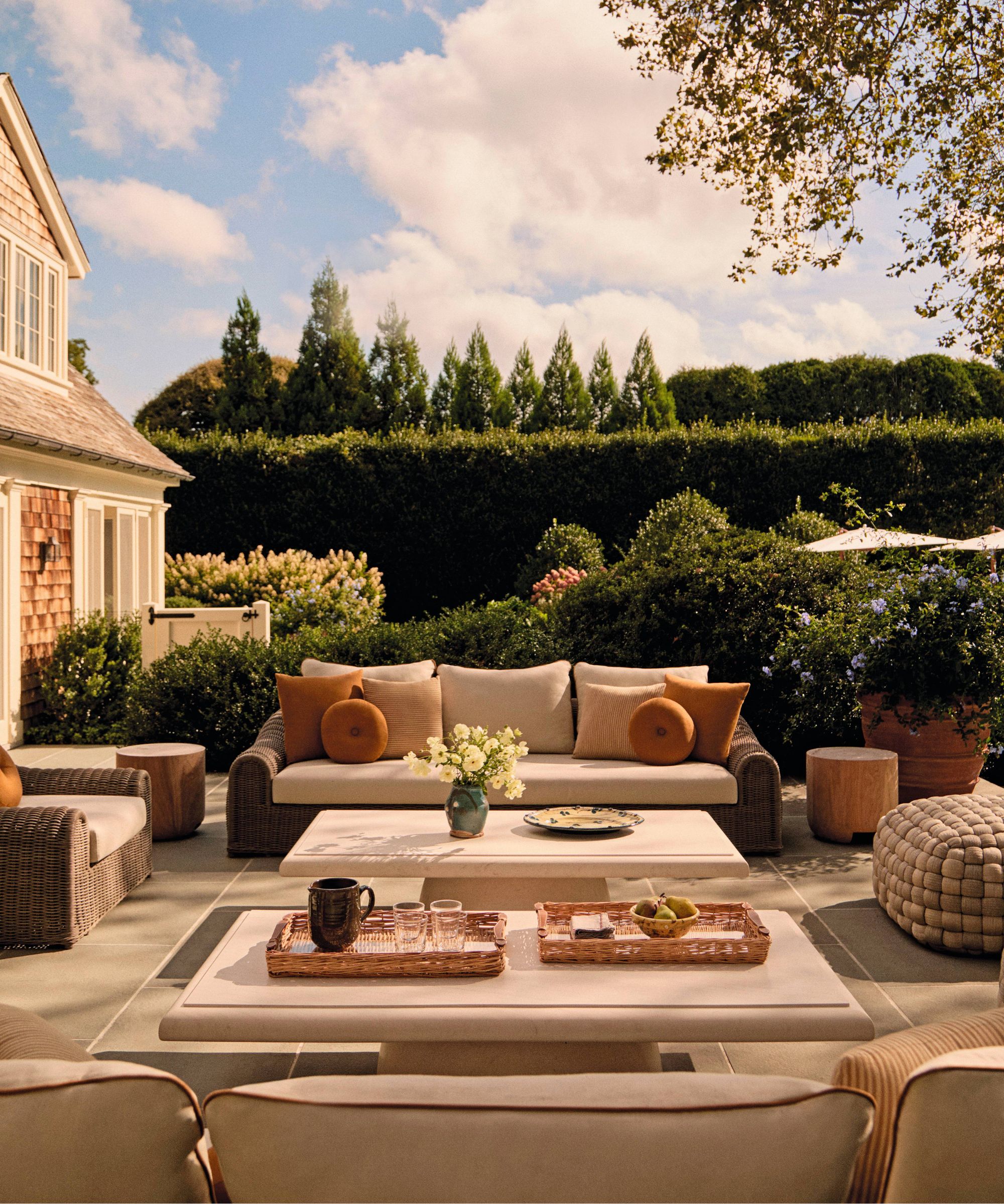 a sunny Hamptons backyard with large hedges and tress and a neutral wicker seating area on a patio with stone coffee tables