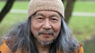 Damo Suzuki modern portrait wearing a crochet hat and orange anorak