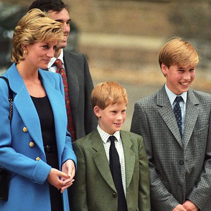 Princess Diana wears a blue suit jacket while posing with young sons Prince Harry and Prince William who wear gray suits during a visit to Eton school