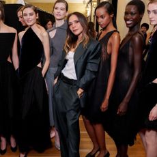 Victoria Beckham poses with models after the Victoria Beckham AW26 runway show wearing dark grey puddle pants with a grey blazer and a white tank top.