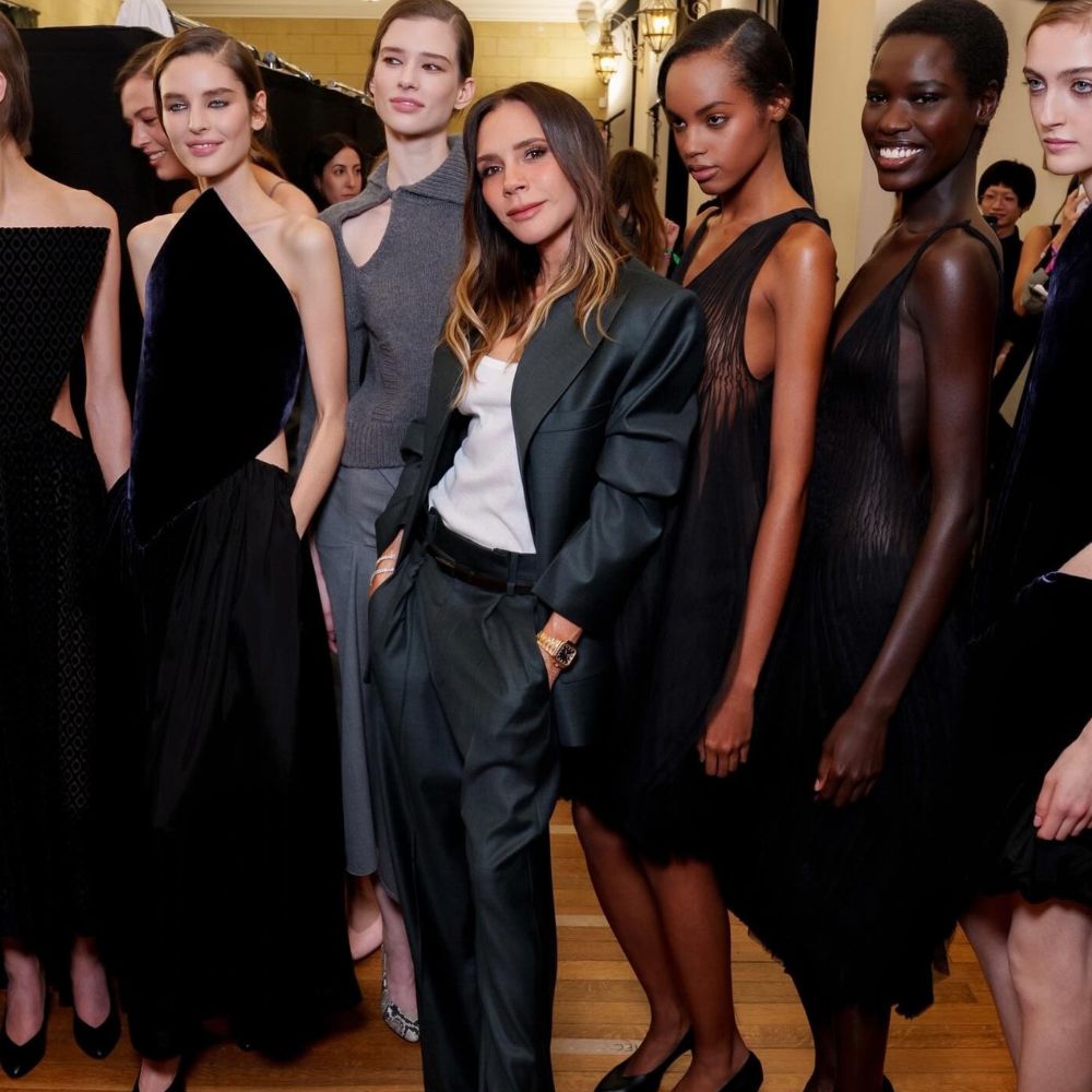 Victoria Beckham poses with models after the Victoria Beckham AW26 runway show wearing dark grey puddle pants with a grey blazer and a white tank top.