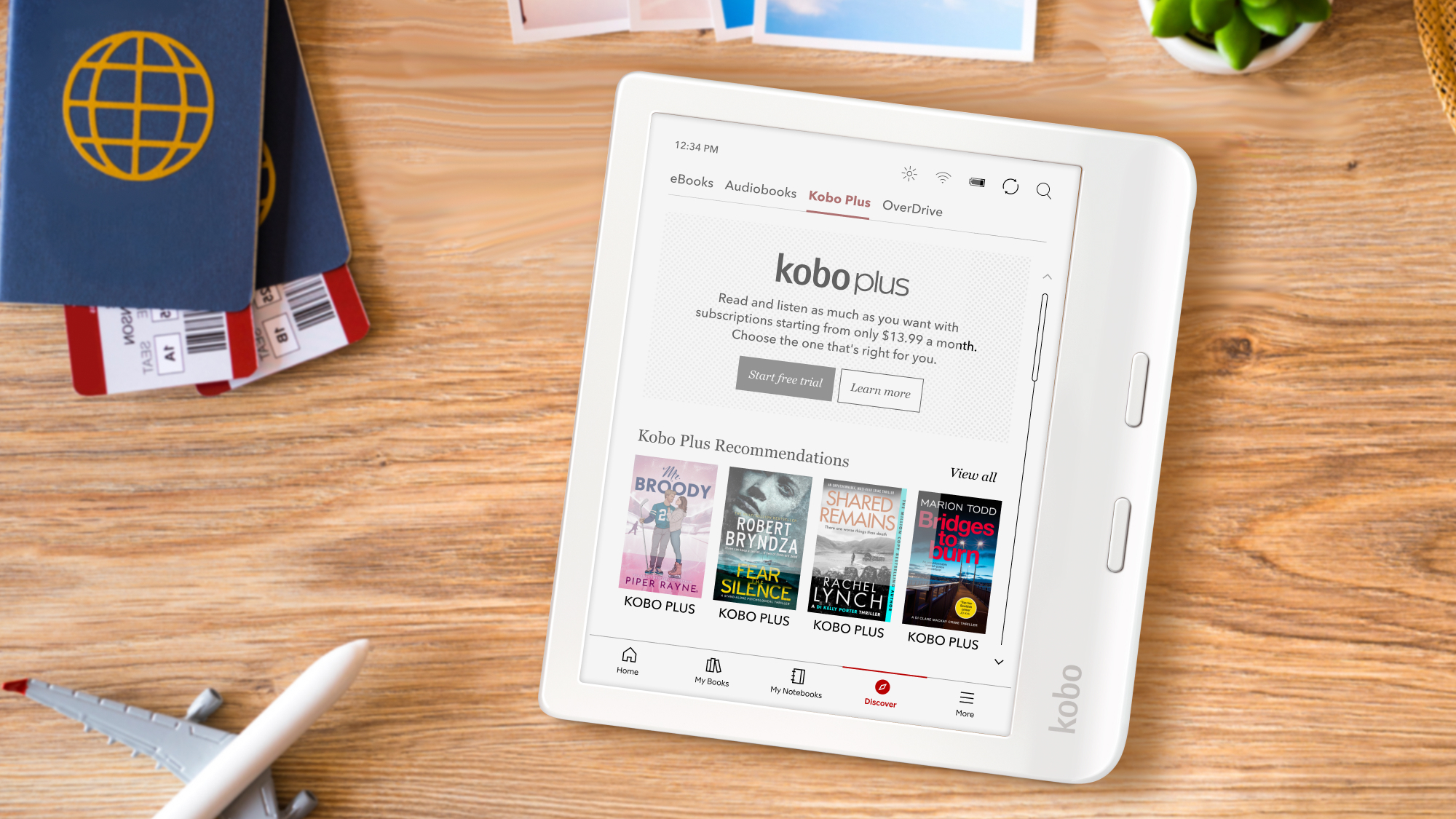 A white digital e-reader displaying kobo plus on a wooden table with a passport and model plane 