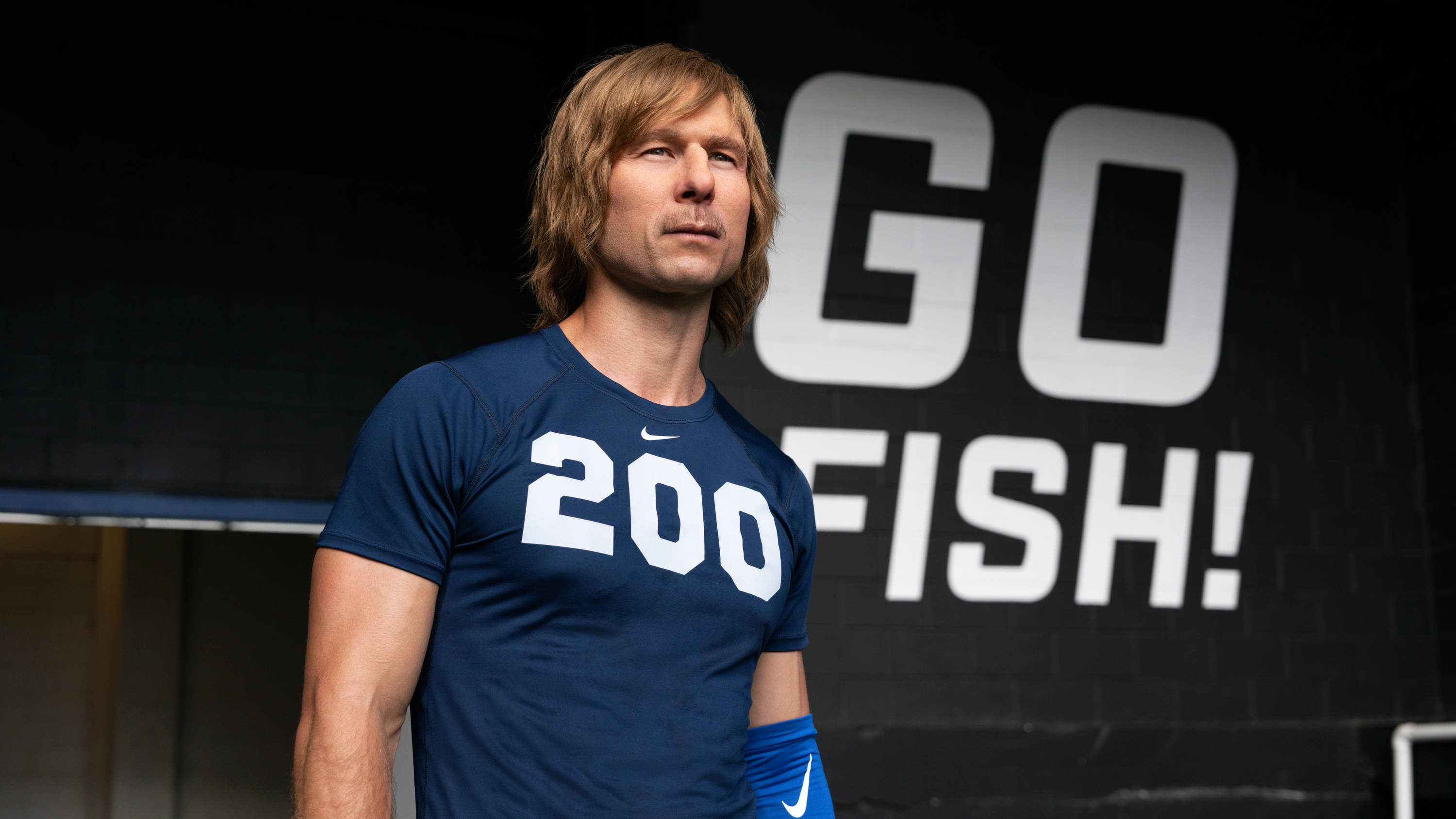 Chad Powers (Glen Powell) standing in front of a "Go Fish!" sign in a scene from Disney/Hulu's "Chad Powers"