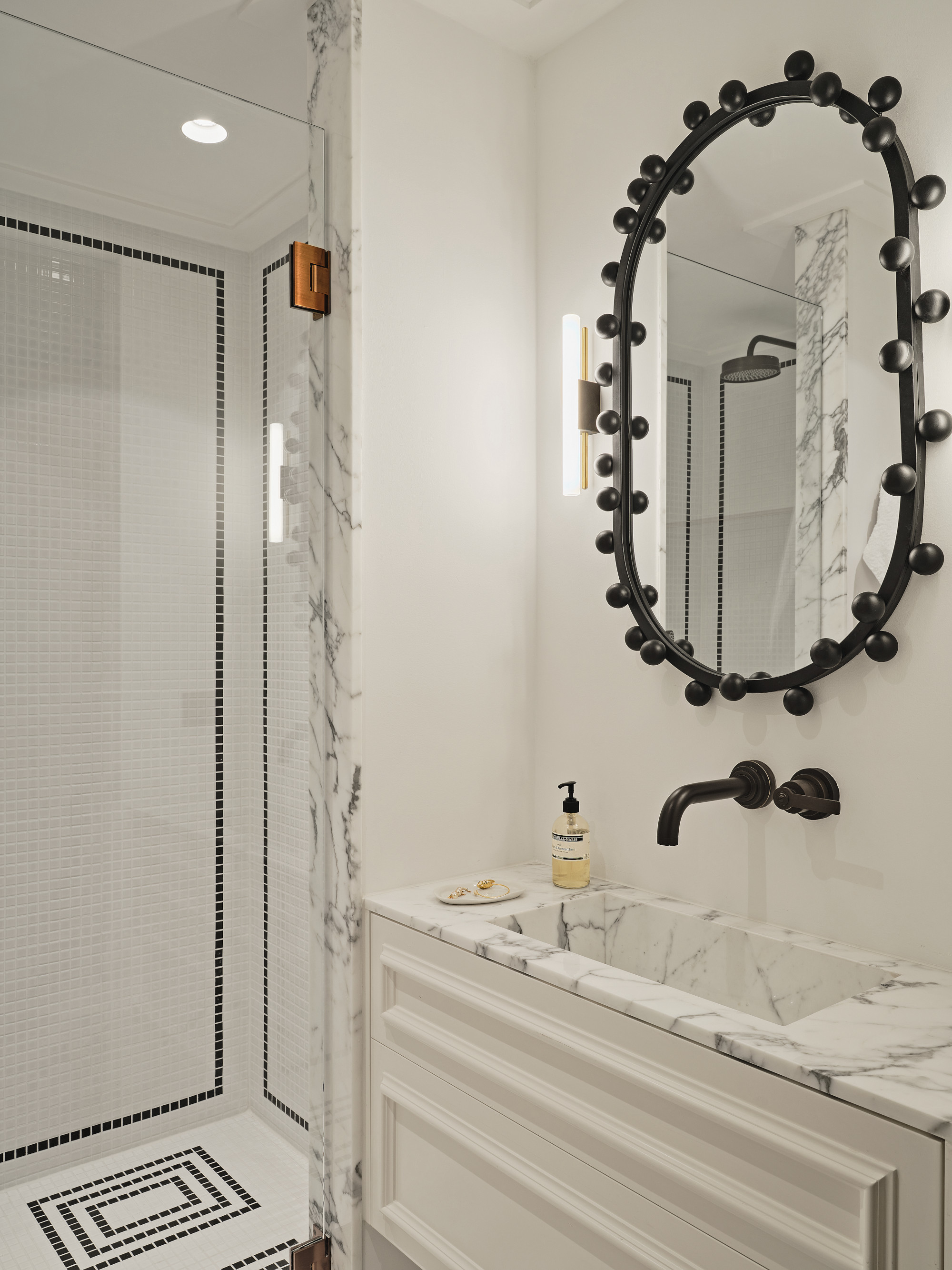 White bathroom with white marble countertop and shower frame, black hardware and black bobble mirror, with white mosaic shower tiles with black linear design