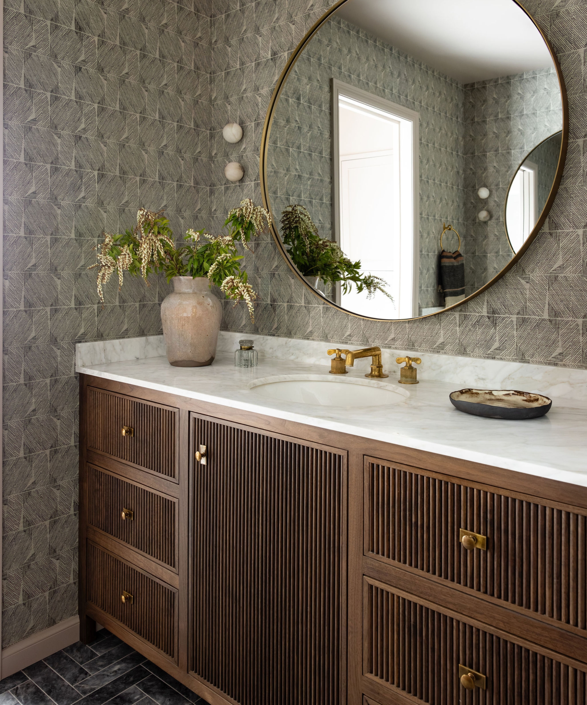 Primary bathroom with a ribbed wood vanity, marble countertop, brass fixtures, round mirror and geometric wallpaper.