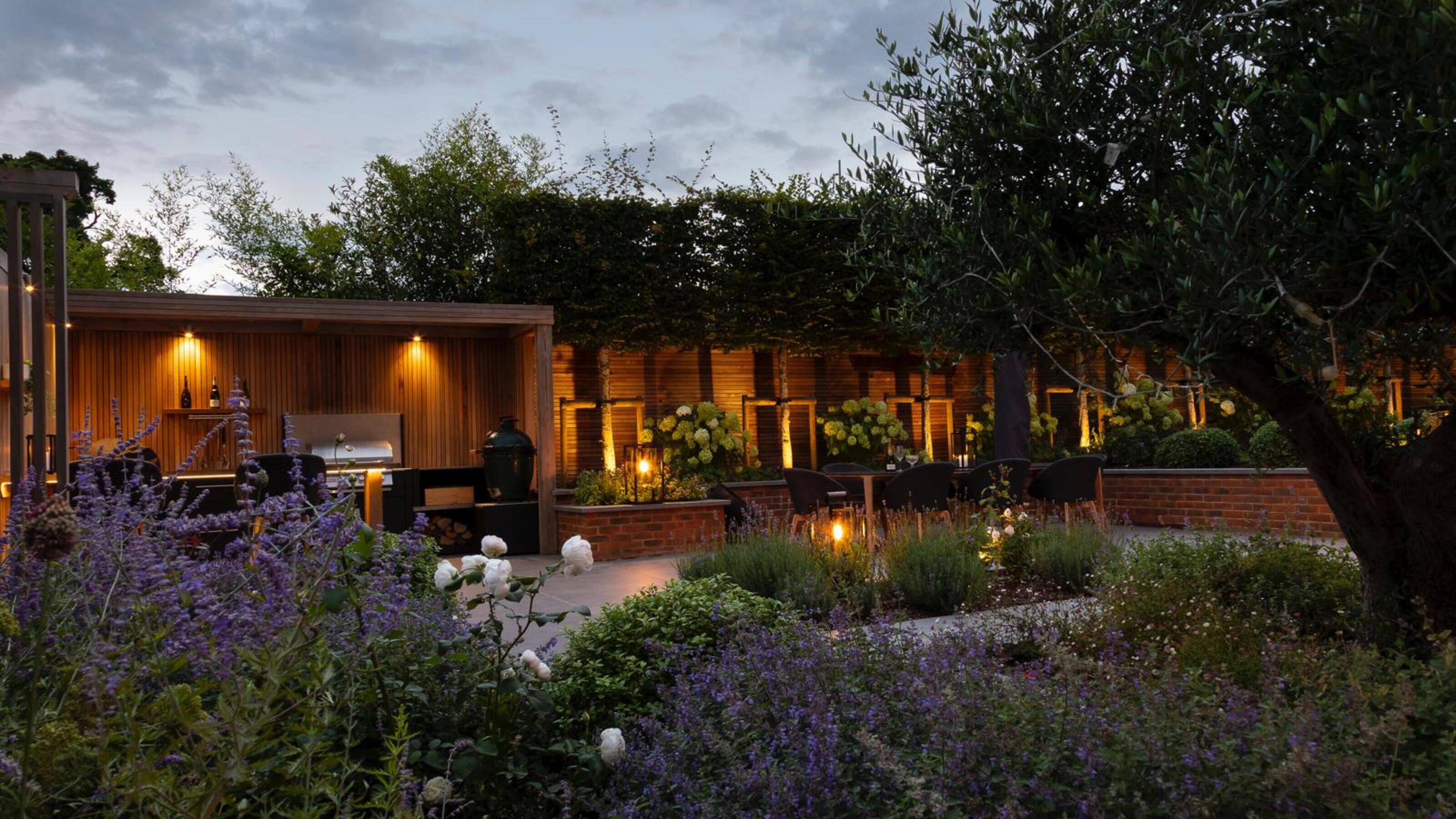 A modern garden with layered planting, an outdoor kitchen, a slatted fence, and uplights lighting a row of trees