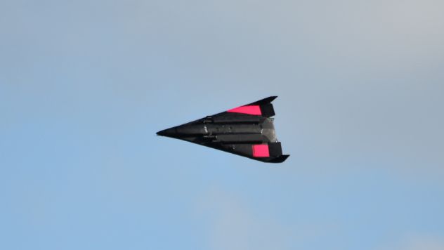 a triangular black plane with two differently-shaped red patches on each wing flies against a blue sky.