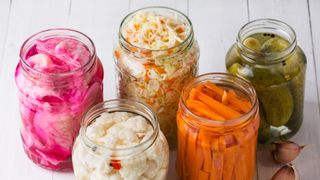 a selection of probiotics, including pickles and kimchi, in glass containers sitting on white wooden table