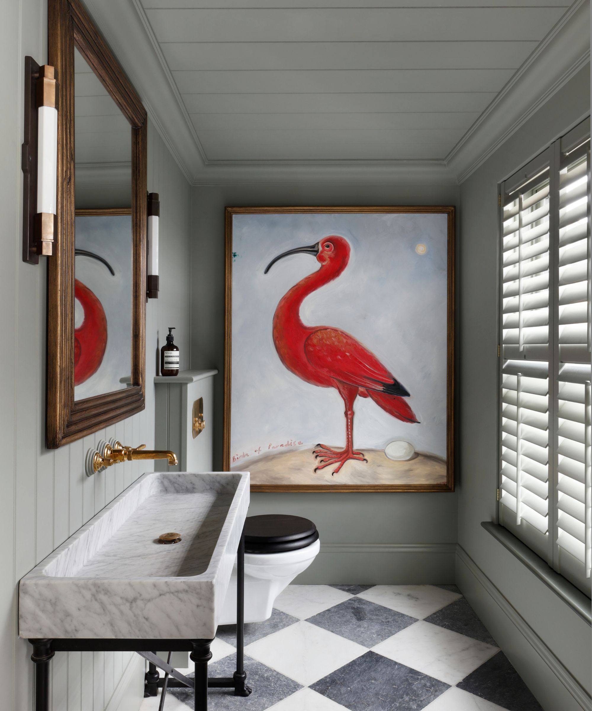 A bathroom with black and white checkerboard floor tiles, gray panelled walls and ceiling, a marble sink with a brass tap, and a large piece of artwork on the wall of a red bird.