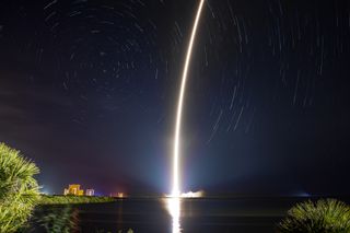 The long exposure streak of a rocket launches against a star-streaked night sky.