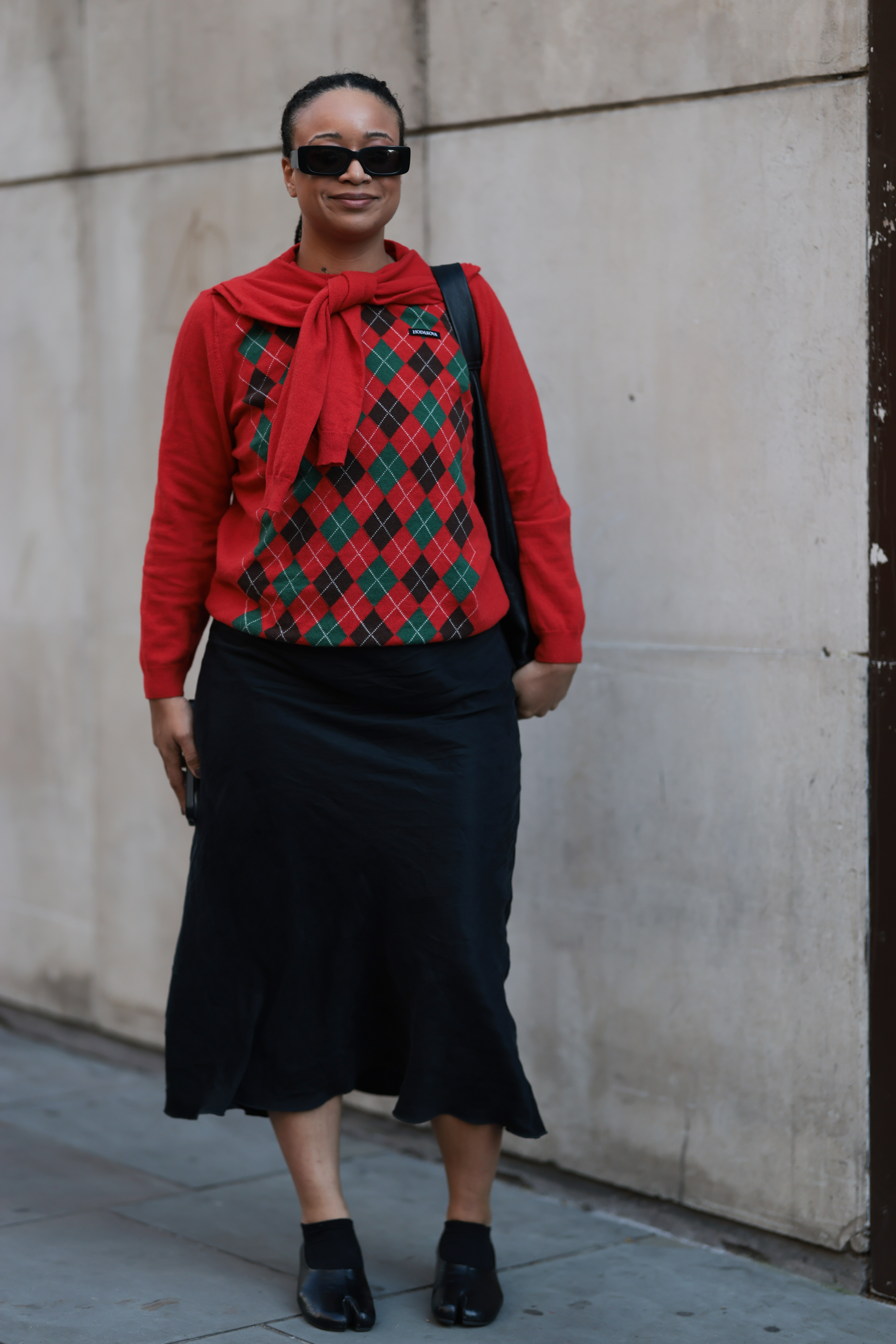 LONDON, ENGLAND - SEPTEMBER 19: Fashion week guest is seen wearing a bright red sweater featuring a green and black argyle pattern across the chest, with a matching red sweater draped around her neck. Her lower half is clad in a long, black, flowing skirt. She accessorizes with black sunglasses and a black shoulder bag, and holds a phone in her left hand. On her feet, she wears black split-toe shoes on Oscar Ouyang Show during London Fashion Week on September 19, 2025 in London, England. (Photo by Jeremy Moeller/Getty Images)