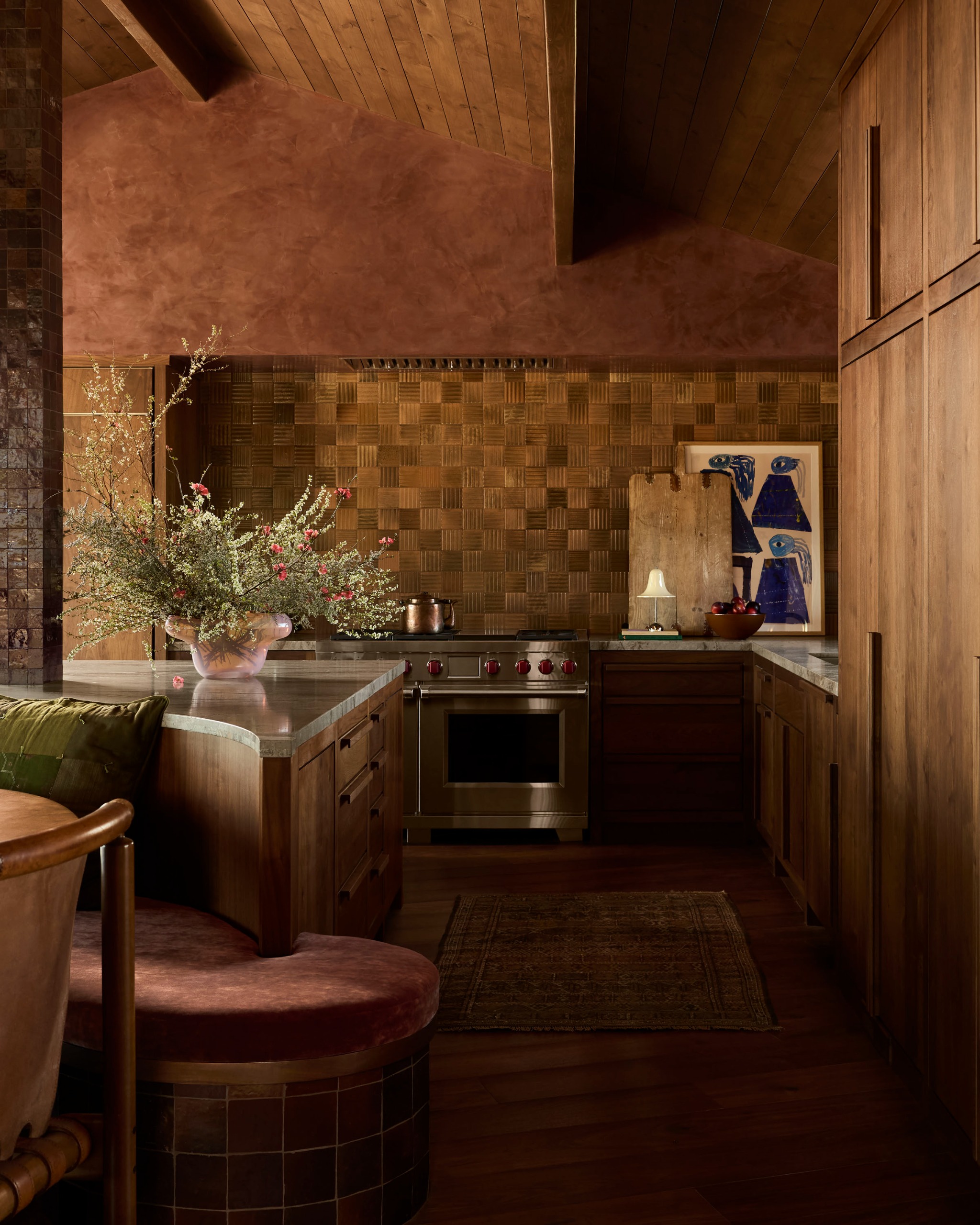 A modern kitchen with an ochre tiled backsplash, wood paneling and cabinetry, marble counters and a custom curved kitchen bench with a large vase of flowers on the kitchen island