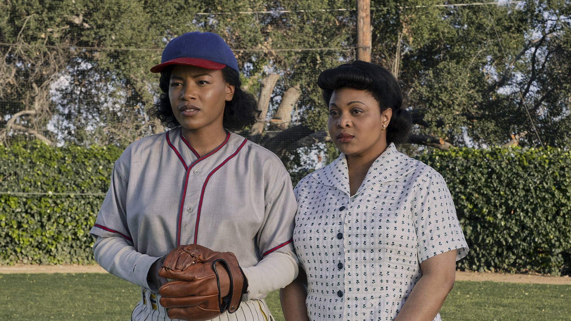 (left to right) Shanti Adams (Max) wears her baseball uniform holding a hood while Gbemisola Ikumelo (Clance) stands next to her holding a briefcase at A League of Their Own